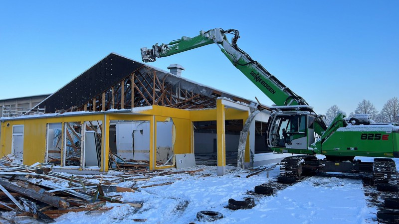 Grüner Abbruchbagger beim kontrollierten Abriss eines gelben Gebäudes im Winter